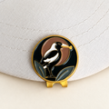 The Course Thief magpie golf ball marker by Fore & Femme clipped to a cream linen cap